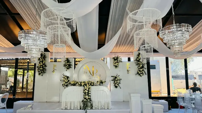 A white-draped wedding reception with crystal chandeliers, floral pillars and a gold monogram backdrop behind the bridal table